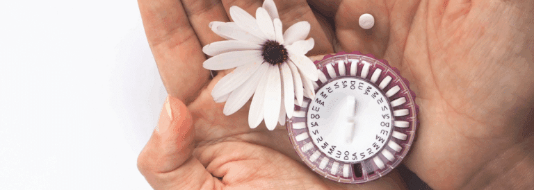 Manos sosteniendo una flor blanca y un envase circular de pastillas anticonceptivas sobre un fondo blanco