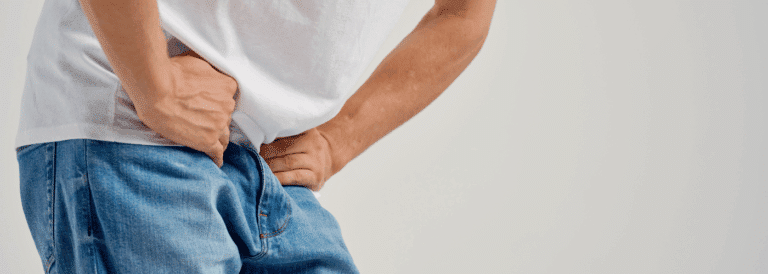Hombre joven con las manos apoyadas en la zona inguinal y el abdomen bajo, inclinado hacia adelante, con gesto de molestia. Viste remera blanca y jeans. Fondo neutro.
