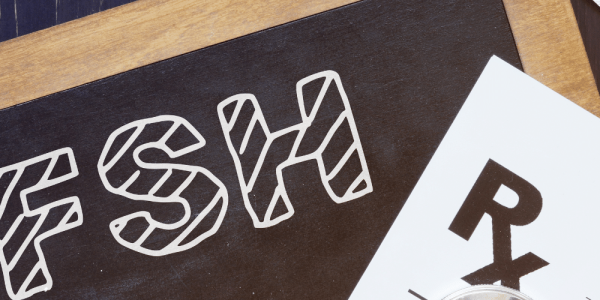 Placa negra con las siglas FSH en tipografía blanca, acompañada de una receta médica y un blíster de comprimidos sobre una superficie de madera.