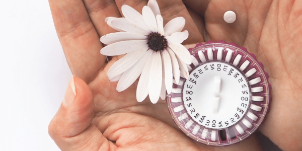 Manos sosteniendo una flor blanca y un envase circular de pastillas anticonceptivas sobre un fondo blanco