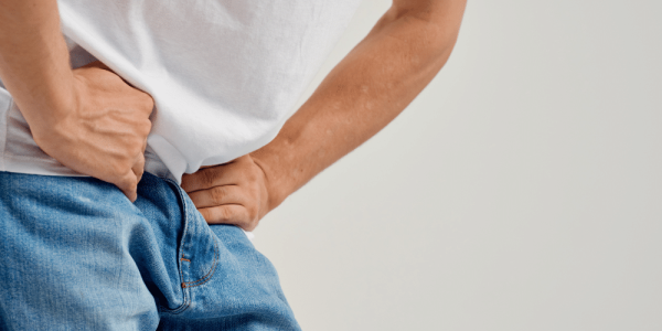 Hombre joven con las manos apoyadas en la zona inguinal y el abdomen bajo, inclinado hacia adelante, con gesto de molestia. Viste remera blanca y jeans. Fondo neutro.